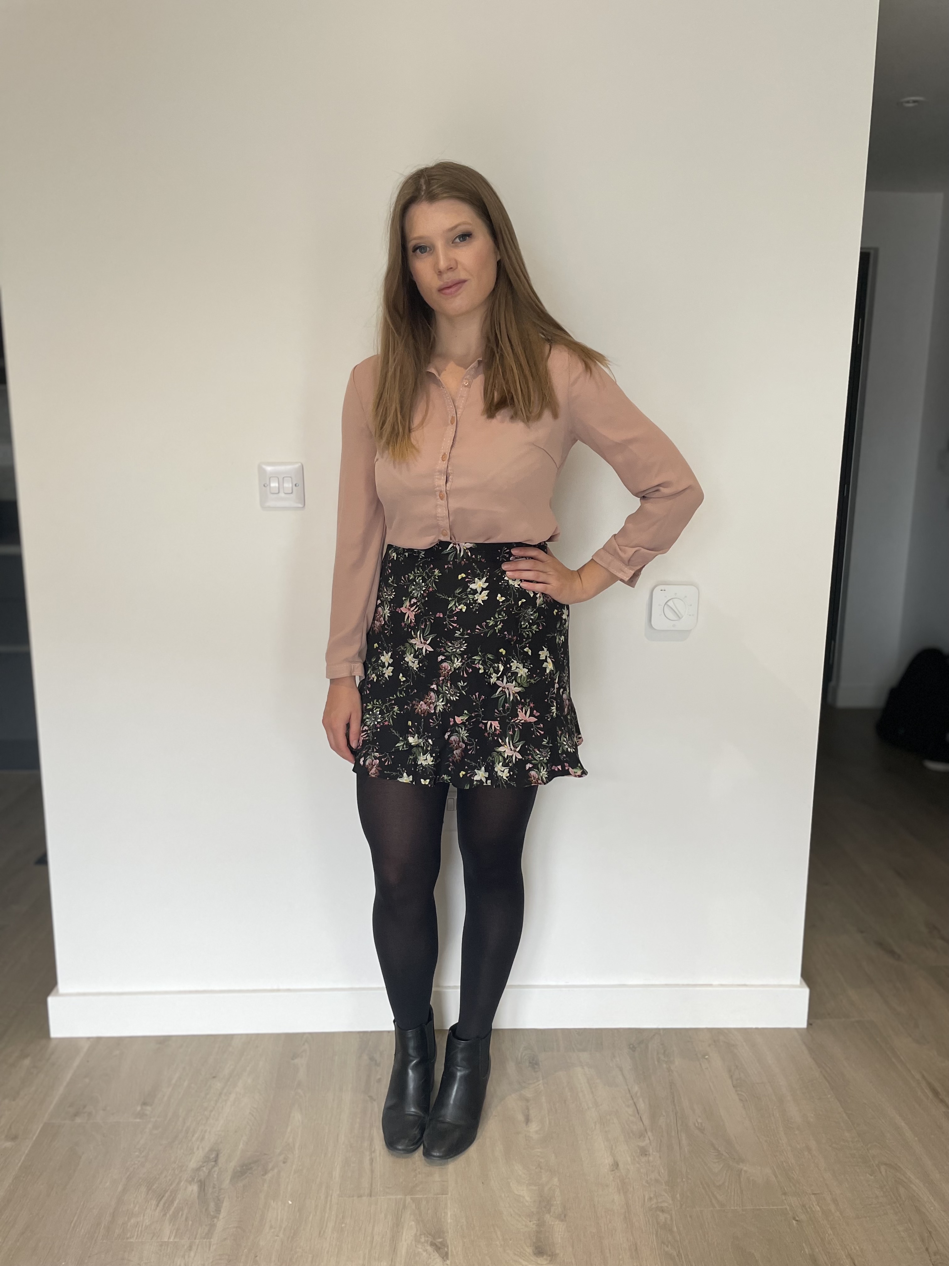 Outfit is a short floral skirt with black tights, black heeled boots and a pale pink shirt.