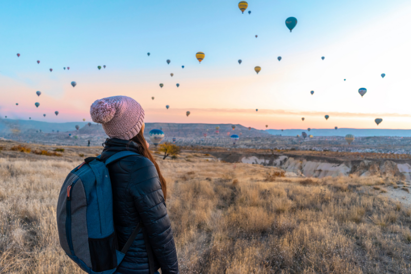 A woman with a backpack stood in the countryside looking at hot air balloons.