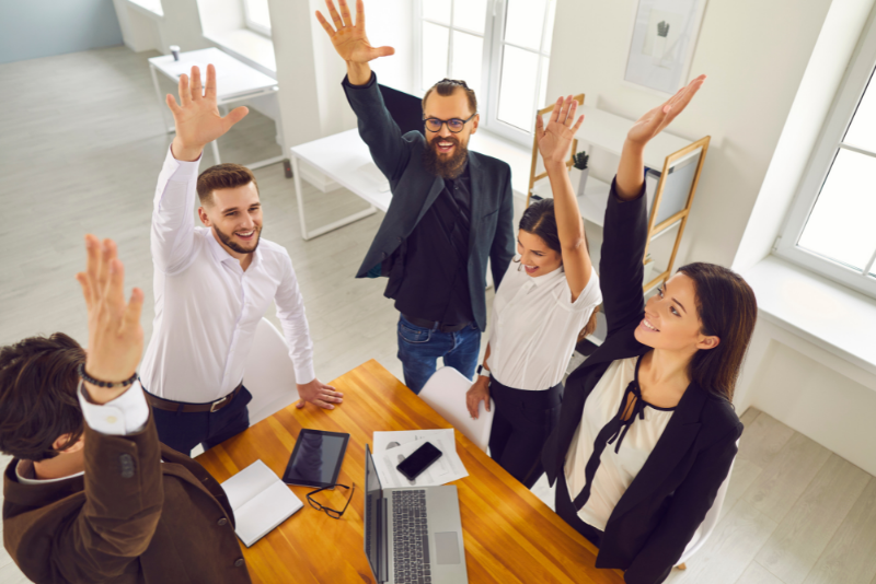 Five work colleagues around a desk raising their hands in the air.